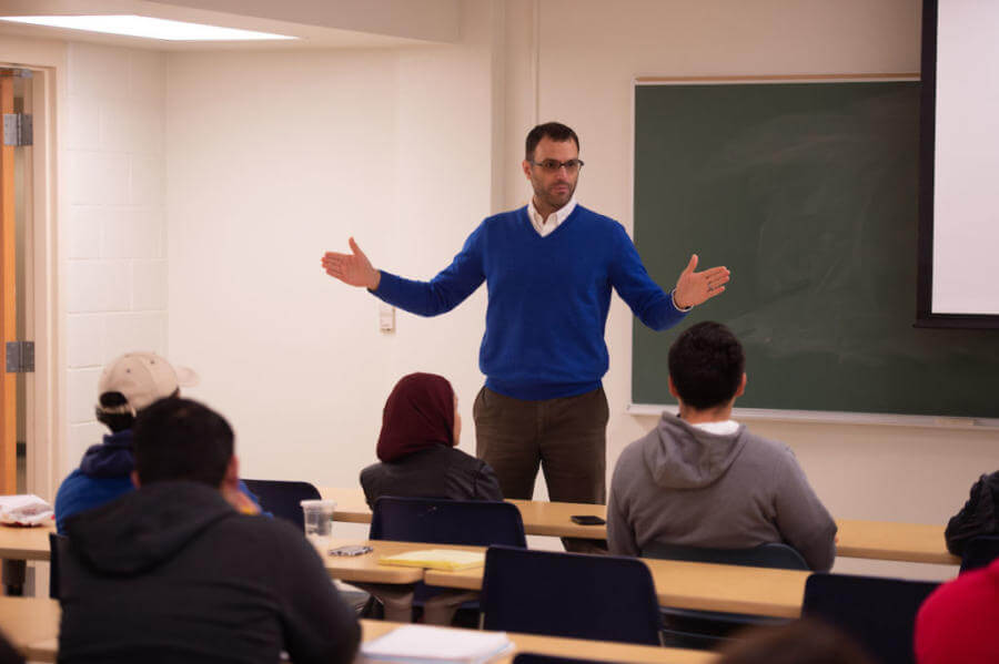 International Studies professor stands in front of a class with his arms spread wide for emphasis