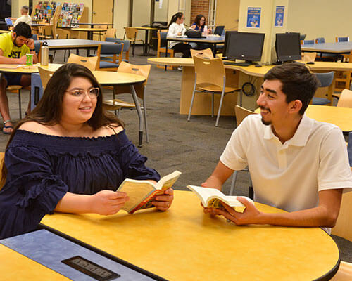 Two students smile and talk while seated at a round table in the library commons.