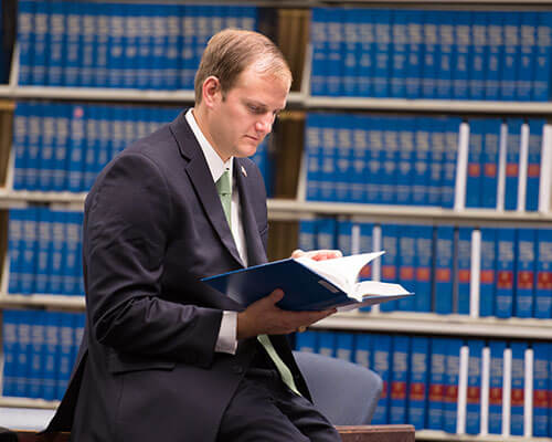 A student in a suit reads a book, with large bookcases full of law books in the background