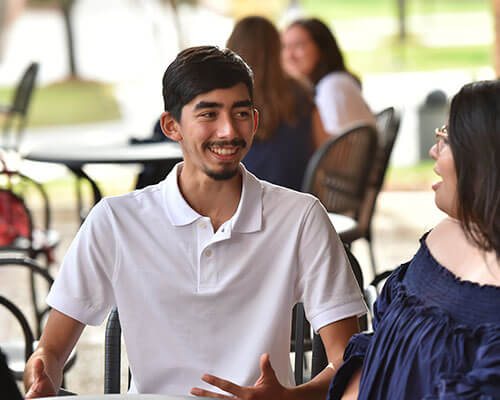 Two students seated outside smile and talk.