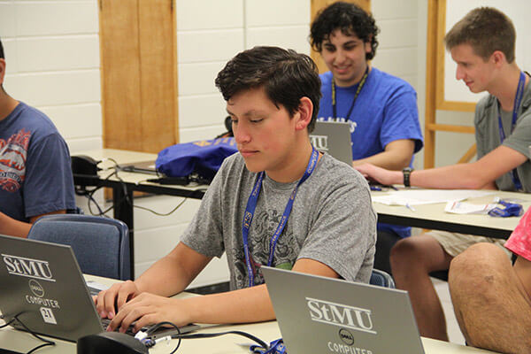 A teenage male student types on a laptop, with other campers in the background
