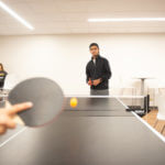 A student's hand and ping-pong paddle blur in the foreground, with focus on the opposing student in the background, playing ping-pong in a lounge in Dougherty Hall