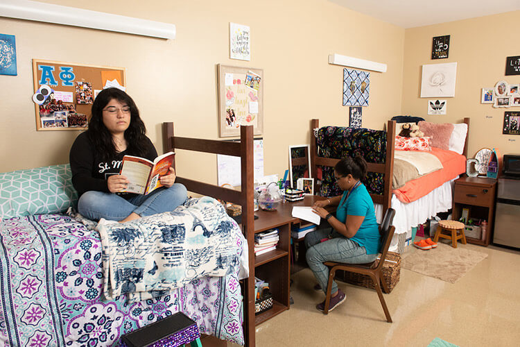 Two female students sit in a Founders Hall room, one on her bed and one reading at a desk