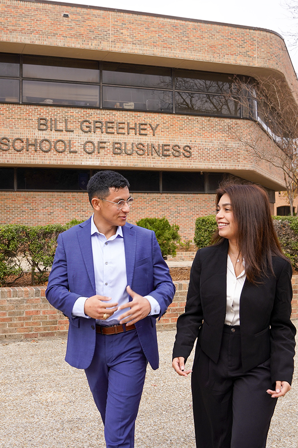 Two students in suits walk and chat in front of the Bill Greehey School of Business. 