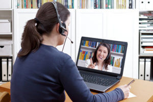 A young woman sits at a computer with a headset on talking to another woman on the computer screen.