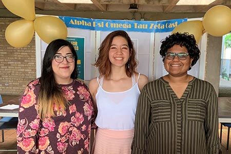 Three female students smiling at a booth promoting Alpha Psi Omega