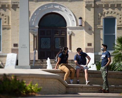 three students with face masks sit in front of St. Louis hall