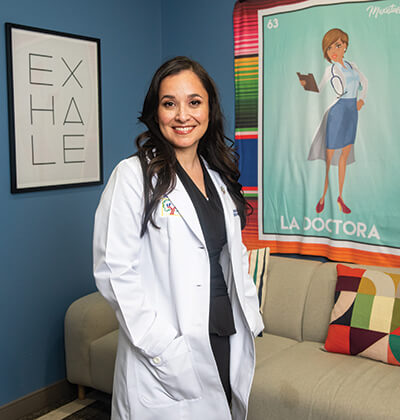 Dr. Erika Gonzalez in her white coat standing in her office with La Doctora tapestry and EXHALE print behind her