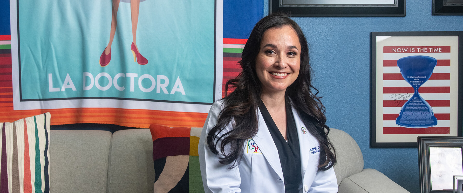 Dr. Erika Gonzalez in her white coat sitting in her office with La Doctora tapestry behind her