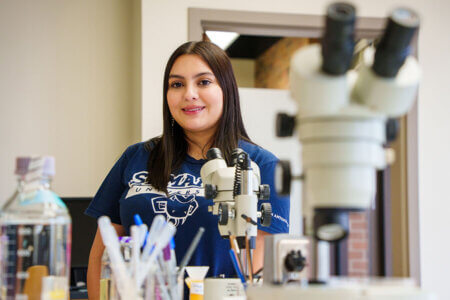 Jocelyn Torres stands near microscopes in a lab.