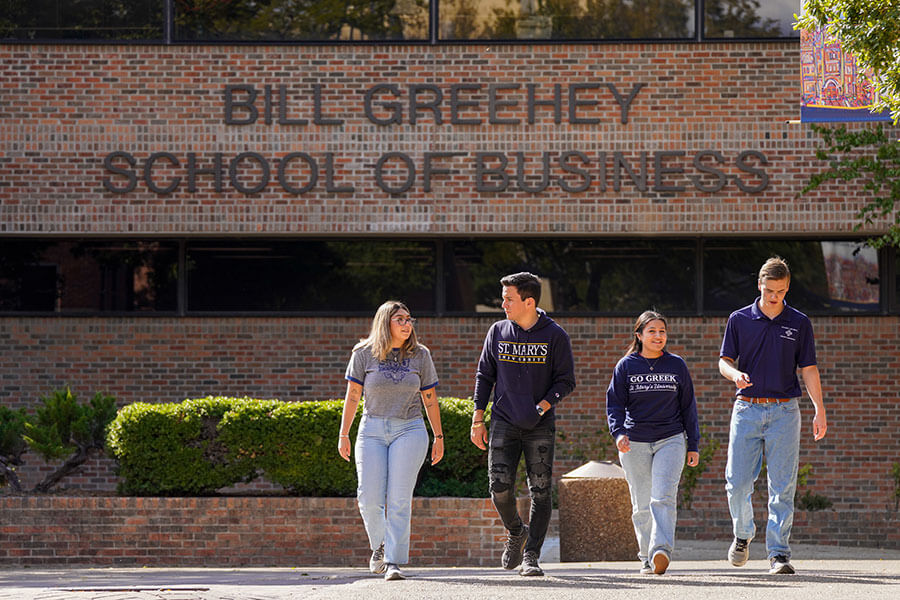 two male and two female students wearing St. Mary's branded attire walk in front of a brick building with the name Bill Greehey School of Business on it
