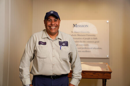 St. Mary's gardener Luis Sanchez smiles in front of a sign with the St. Mary's mission while wearing his work uniform