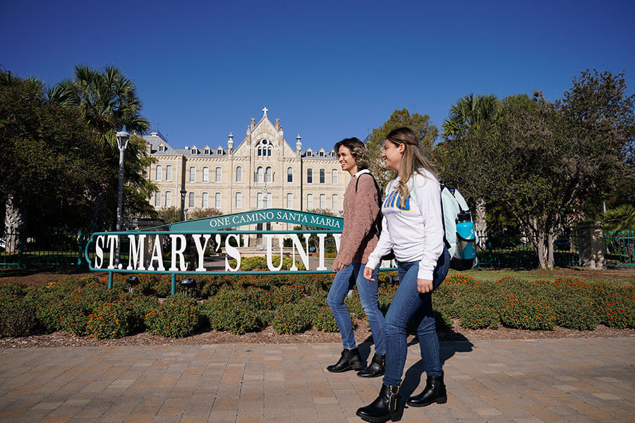 two students with backpacks walk in front of a sign that reads St. Mary's University with St. Louis Hall in the background