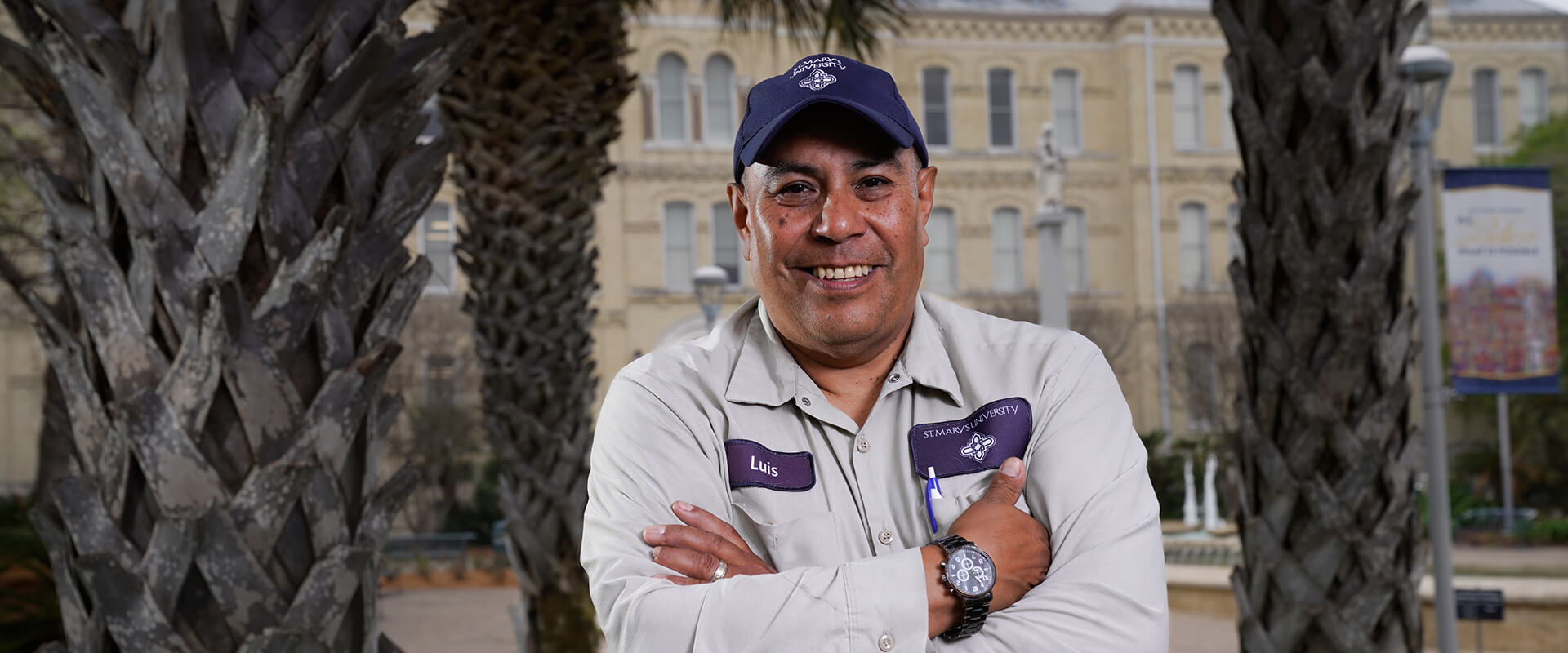 St. Mary's gardener Luis Sanchez smiles in front St. Louis Hall while wearing his work uniform