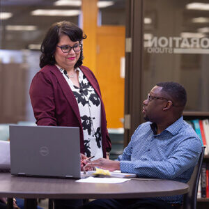 A female faculty member stands by the table where a male student gestures towards his laptop.