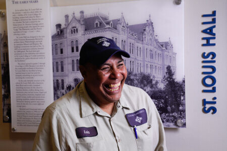St. Mary's gardener Luis Sanchez smiles in front of a sign with a photo of St. Louis Hall while wearing his work uniform