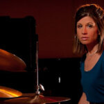 Audra Menconi is seen sitting at a drum kit in a provided photo.