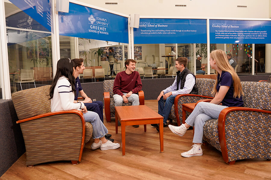 Five students sit on couches in Alkek Building surrounded by signs for Greehey School of Business