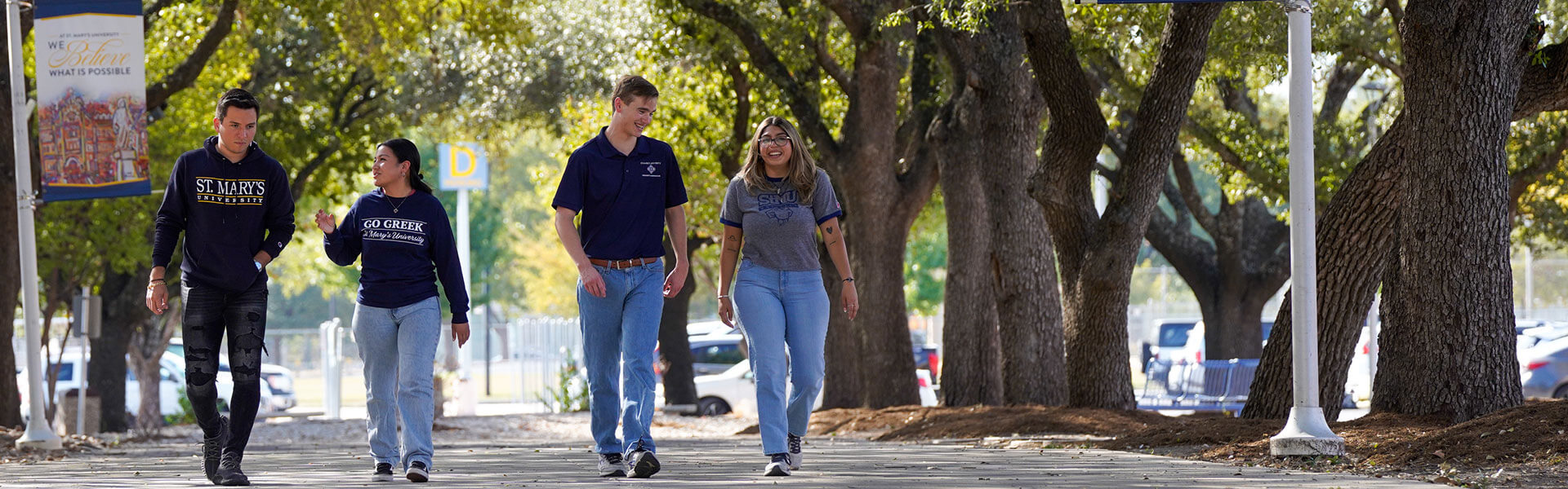 Four students in St. Mary's apparel chat while walking on a tree-lined pathway on campus