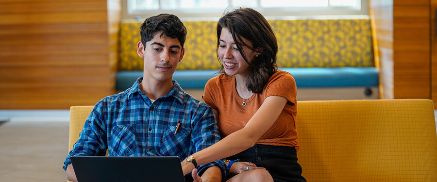 Two students, seated on a brightly colored sofa, study together.