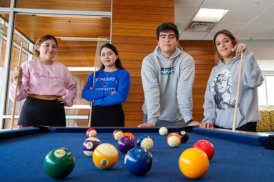 Four students wearing St. Mary's attire pose at a pool table in residence hall lobby.