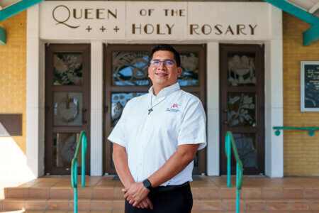 Guillermo “Memo” Peña Contreras posing in Marianist shirt in front of a chapel with the words "Queen of the Holy Rosary" written above the door