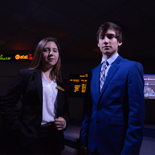 two students stand in front of a stock ticker in a dark room