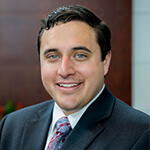 Portrait of Aaron Valadez in a dark suit, white shirt and tie. 