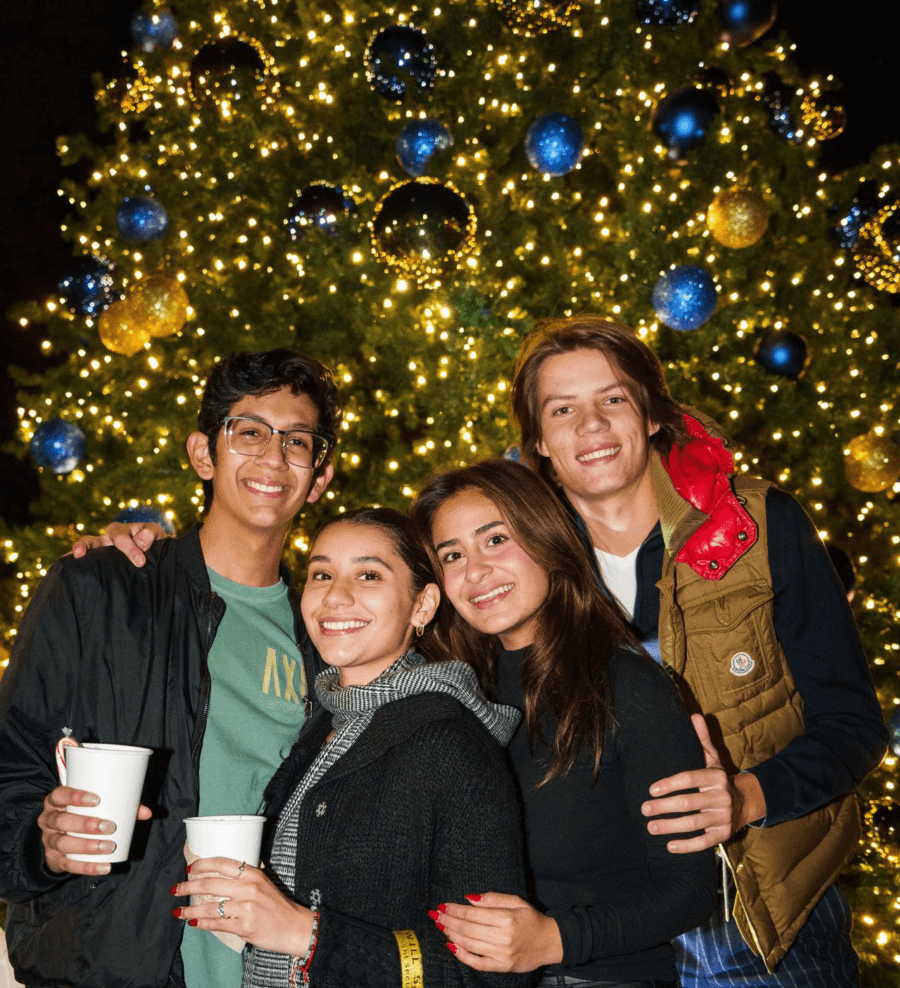 Four students standing in front of a brightly lit Christmas tree holding hot cocoa.