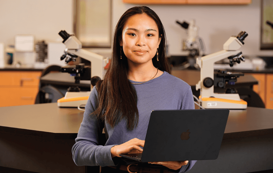 Female student wearing gray long sleeve shirt holds her computer in one hand while smiling. Behind her is the St. Mary's science lab.