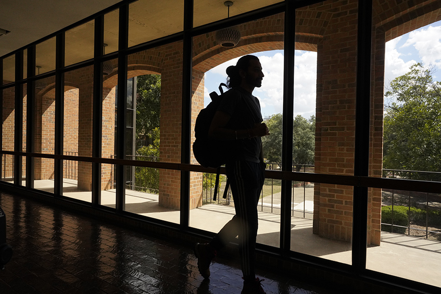 A student walks along a hallway with large windows in the back overlooking a courtyard.