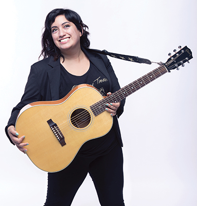 Noosa Al-Sarraj is dressed in all black and holding her guitar.