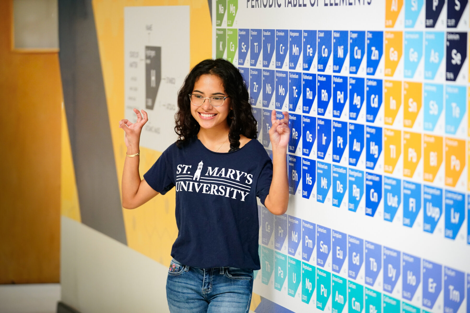 A female wearing a dark blue St. Mary's University shirt shows her Rattler Pride by making the hand gesture Fangs Out in front of a large print out of the Periodic Table of Elements in Garni Hall.