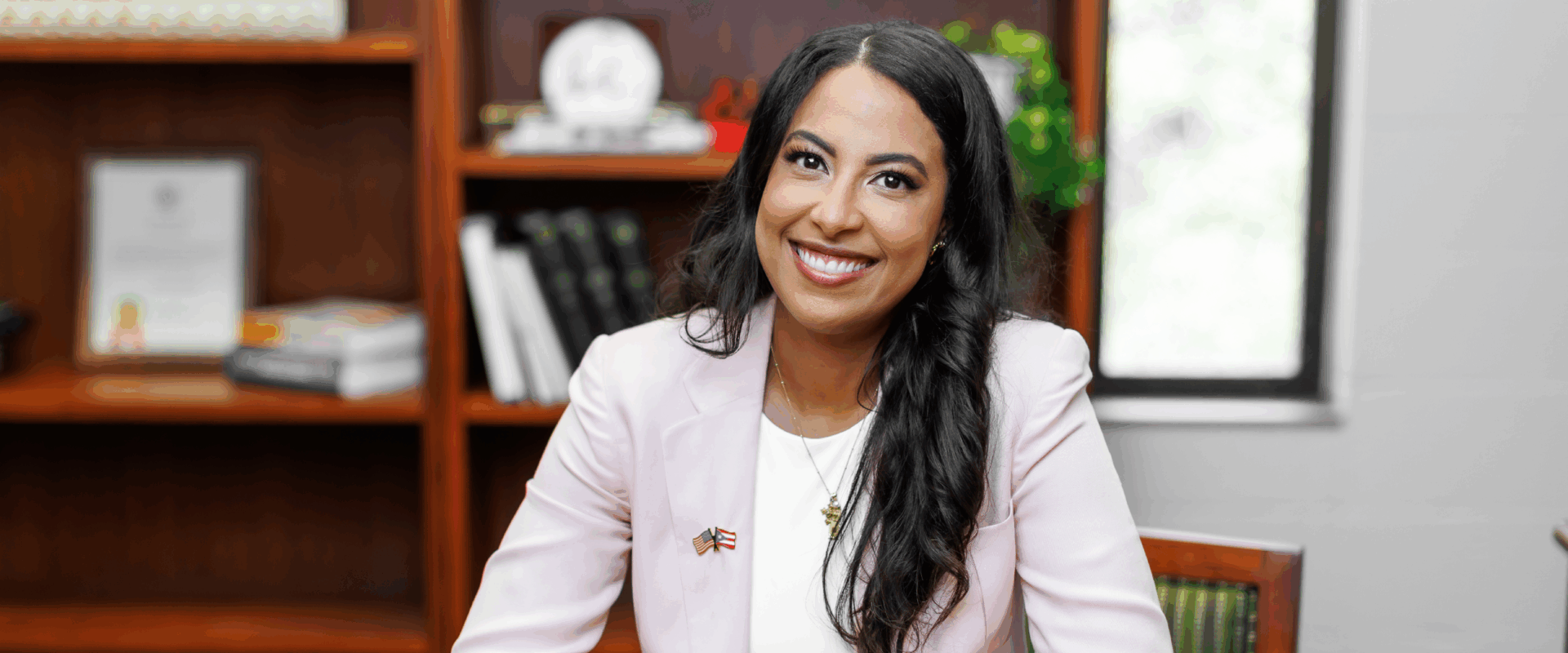 Sigrid Vendrell-Polanco is wearing a pink jacket and white blouse and is seated at her office desk.
