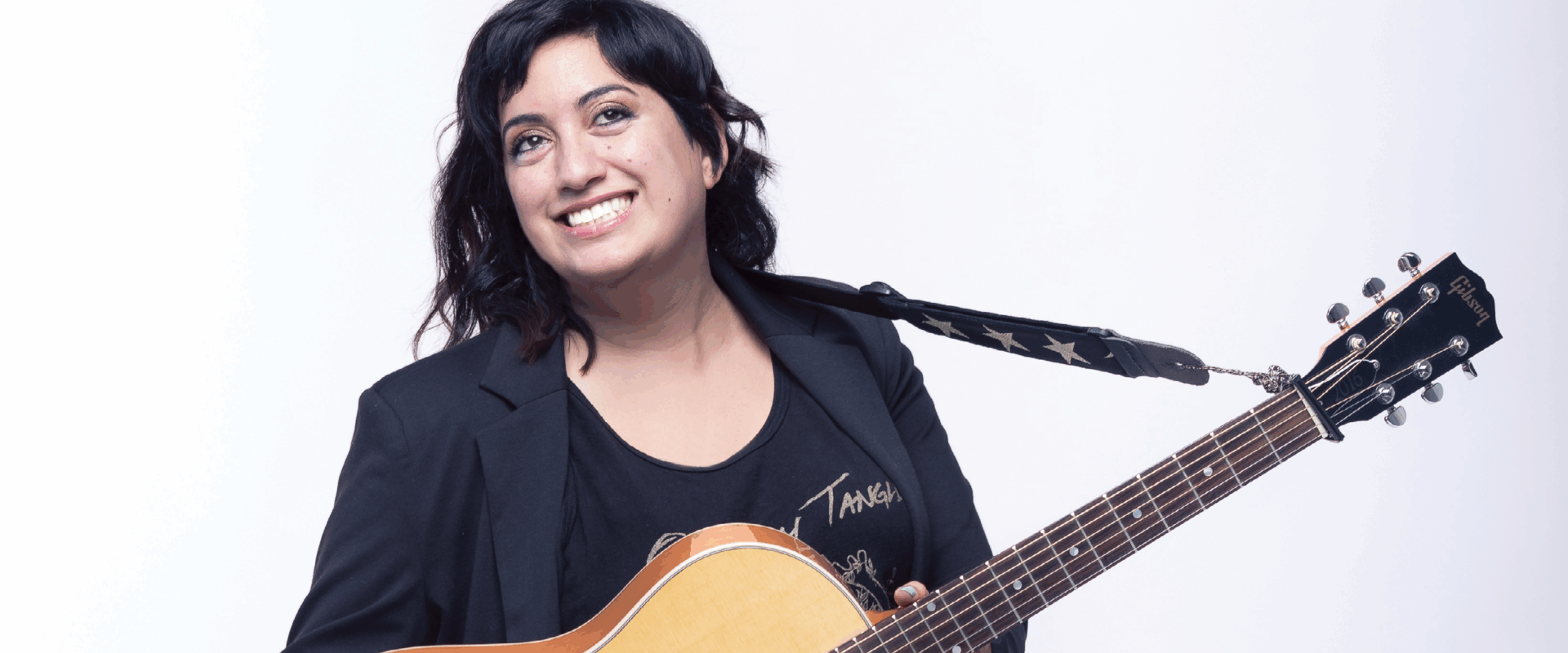 Noosa Al-Sarraj is dressed in all black and holding her guitar.