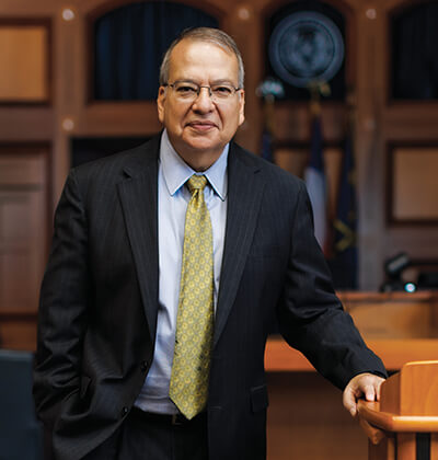 The Hon. Xavier Rodriguez, J.D., in a dark suit standing in the courtroom classroom on campus.