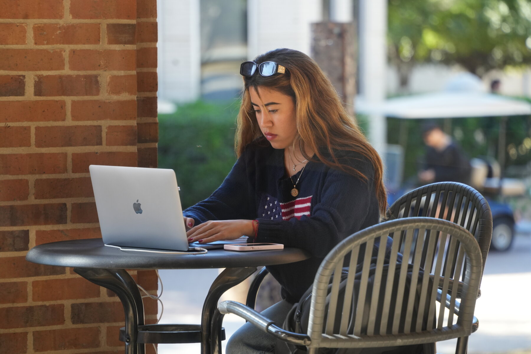 Female student with a United States flag on her shirt enjoys the outdoor weather while using her laptop.