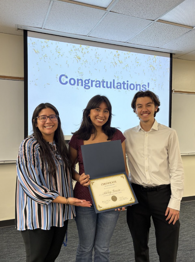 Three students holding a certificate in front of a projector with the projection saying "Congratulations!".