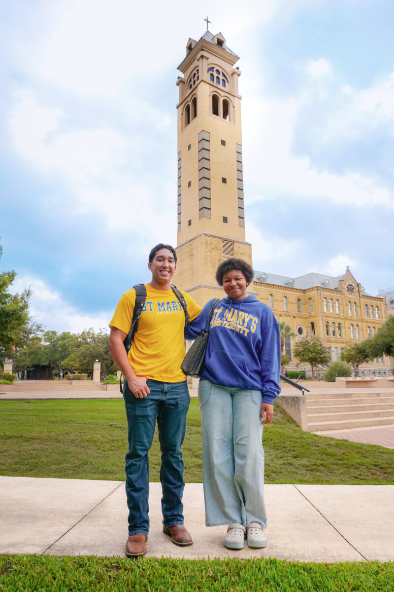 Two students smile and pose for the camera in front of the Barrett Memorial Bell Tower at St. Mary's University.