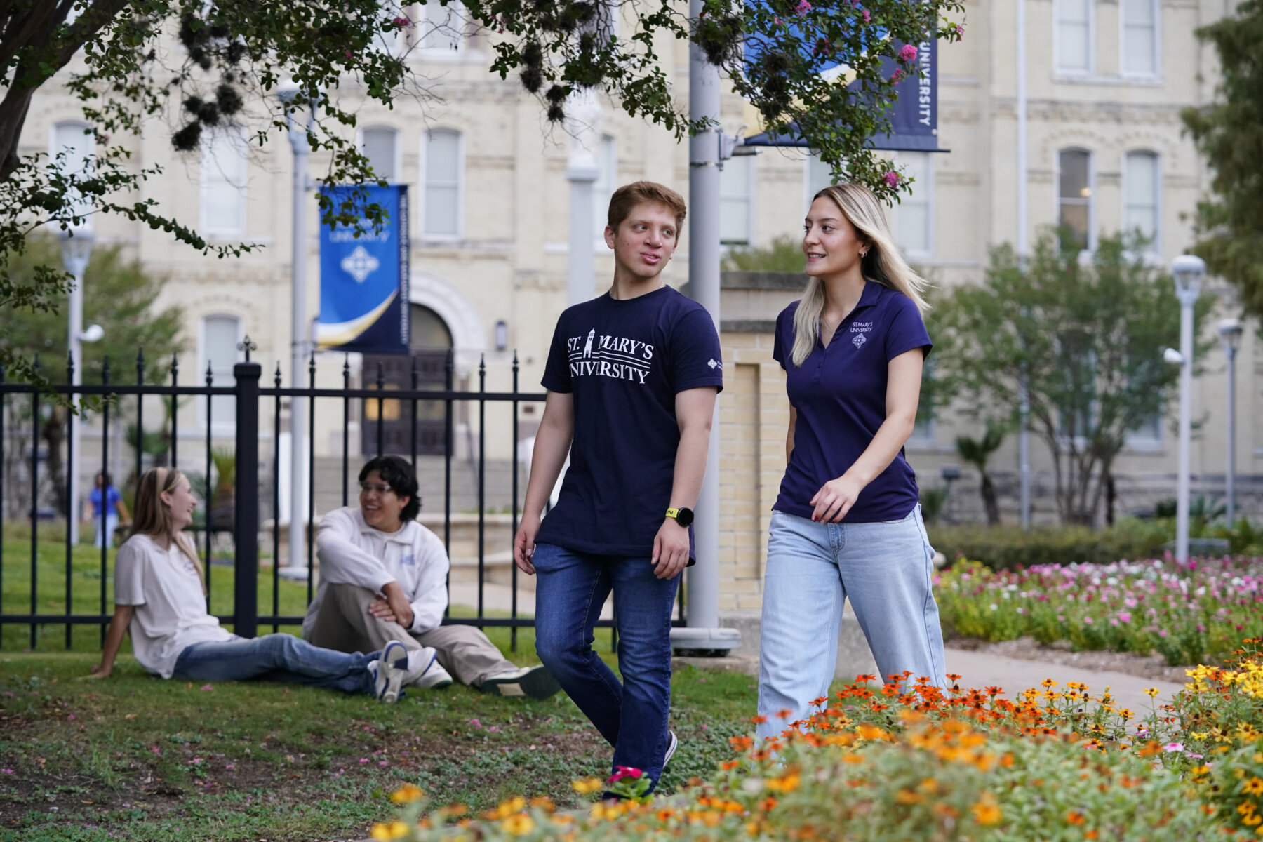 Two students wearing St. Mary's University branded shirts walk by the historic St. Louis Hall. Two other students smile as they converse with each other in the background.