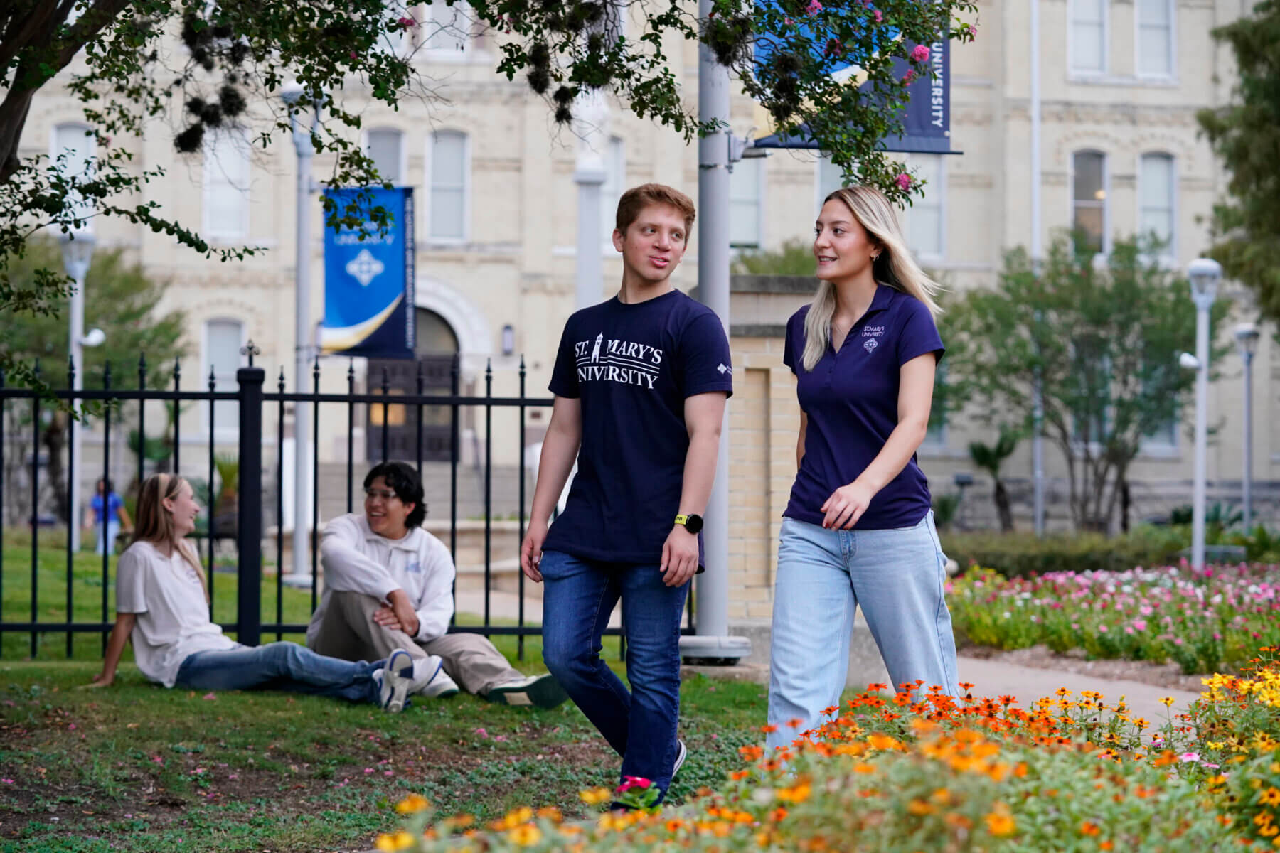 Two students walk on a flower-lined path in front of St. Louis Hall while two students sit on the grass in the background.