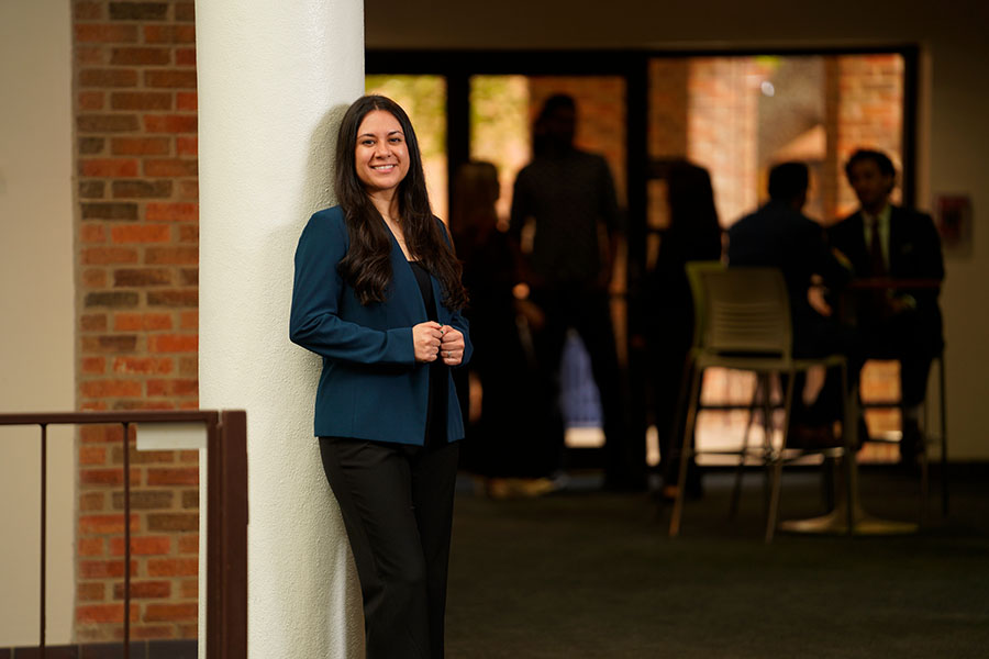 A graduate students smiles while leaning against a pole.