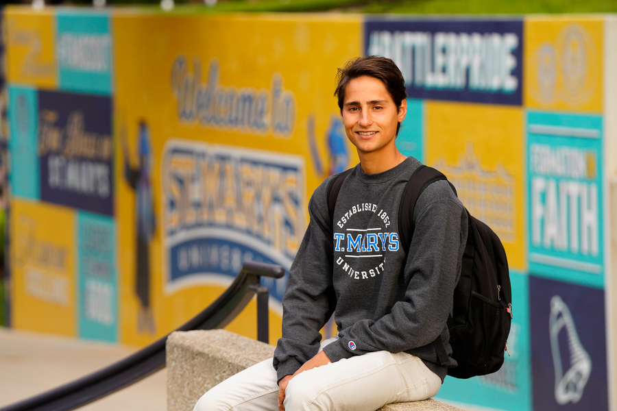 An international St. Mary's student smiles while sitting in front of a gold and blue St. Mary's themed wall.