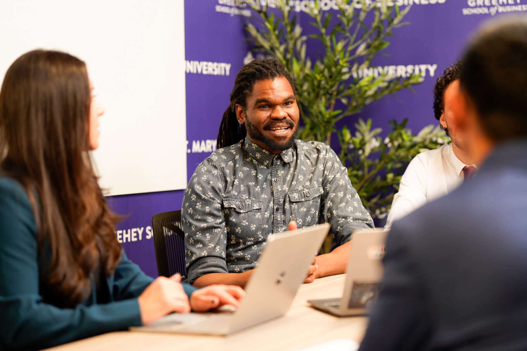A group of students sit around a table having a discussion. On the table are laptops and there is a presentation board on the wall behind one of the students.