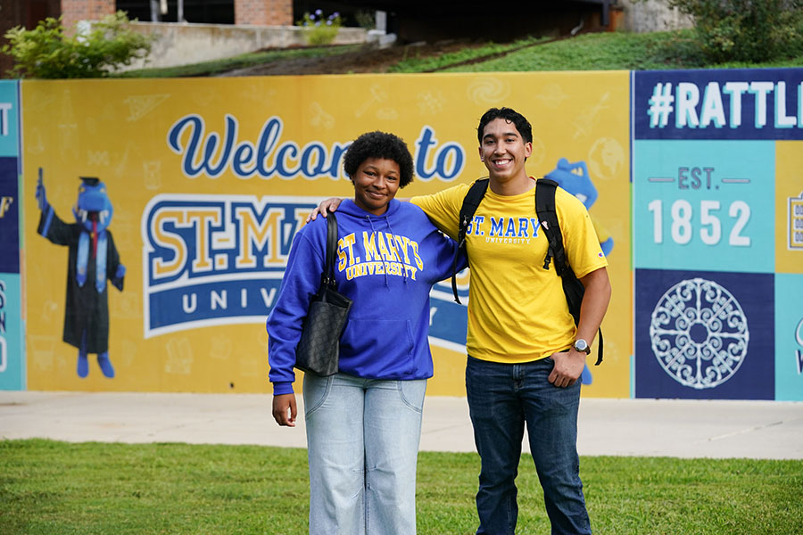 Two students stand with their arms around each other's shoulders in front of a blue and gold St. Mary's themed wall.