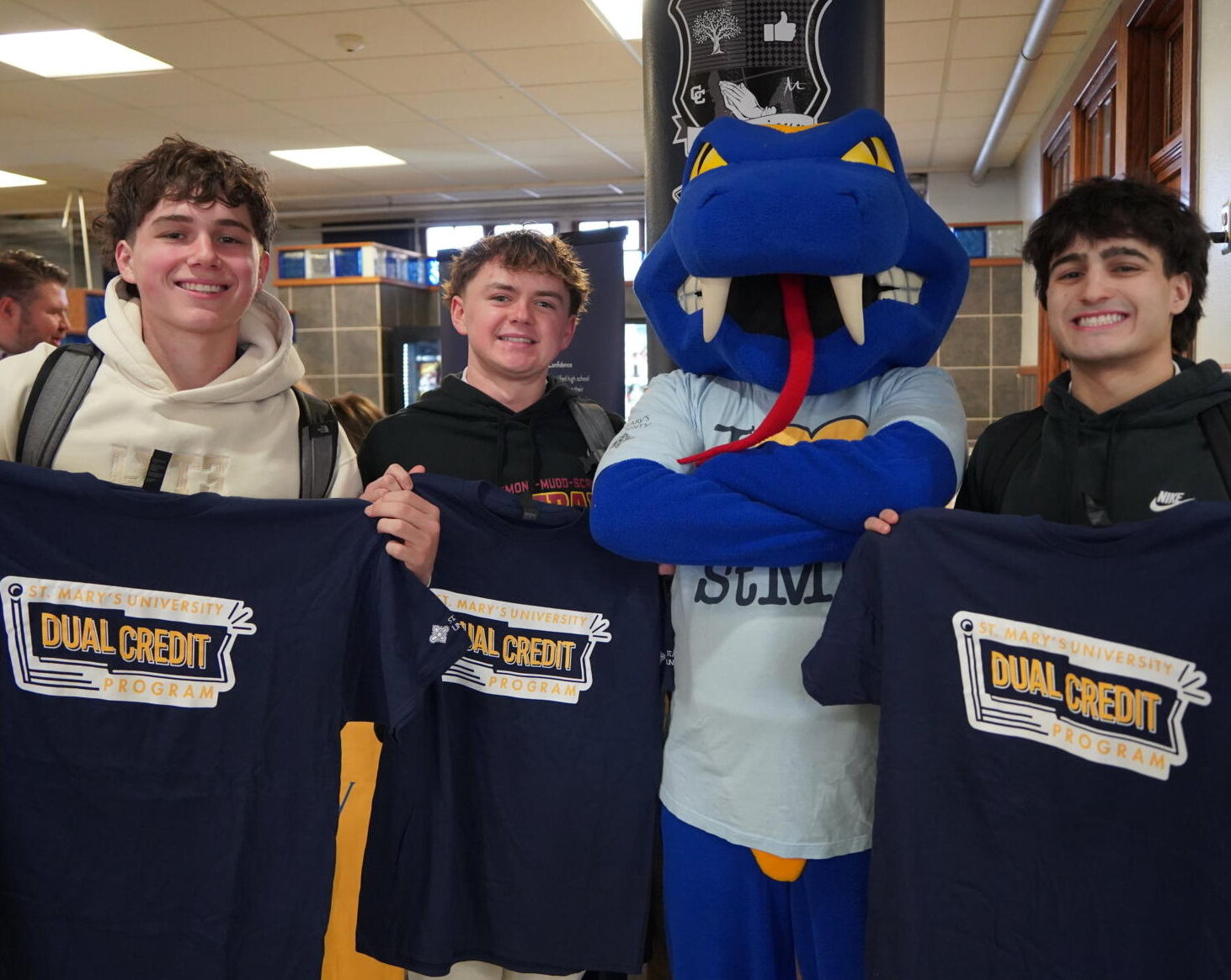 A group of high school boys from Central Catholic hold up their dual credit t-shirts and gather around the St. Mary's mascot, Rattler Man.