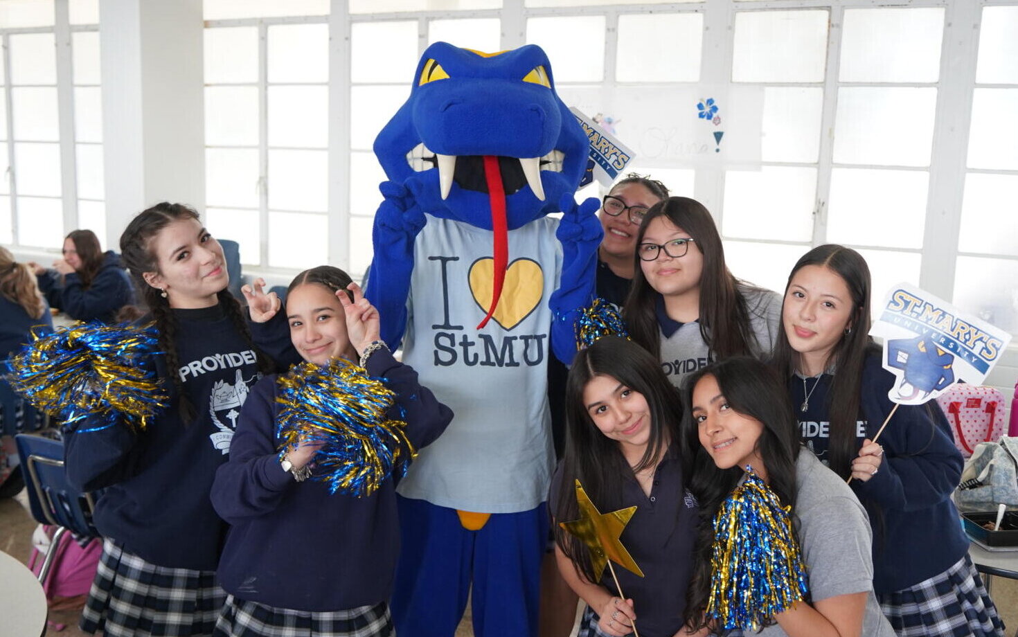 A group of female high school students from Providence gather together around Rattler Man to take a fun photo. They wave pom poms and a St. Mary's University sign in the air with enthusiasm.