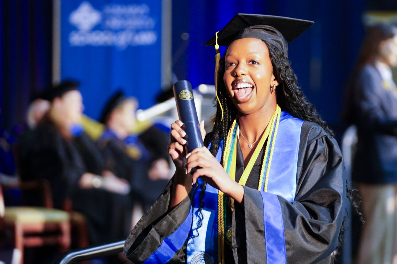 Female student smiles as she holds up her diploma.
