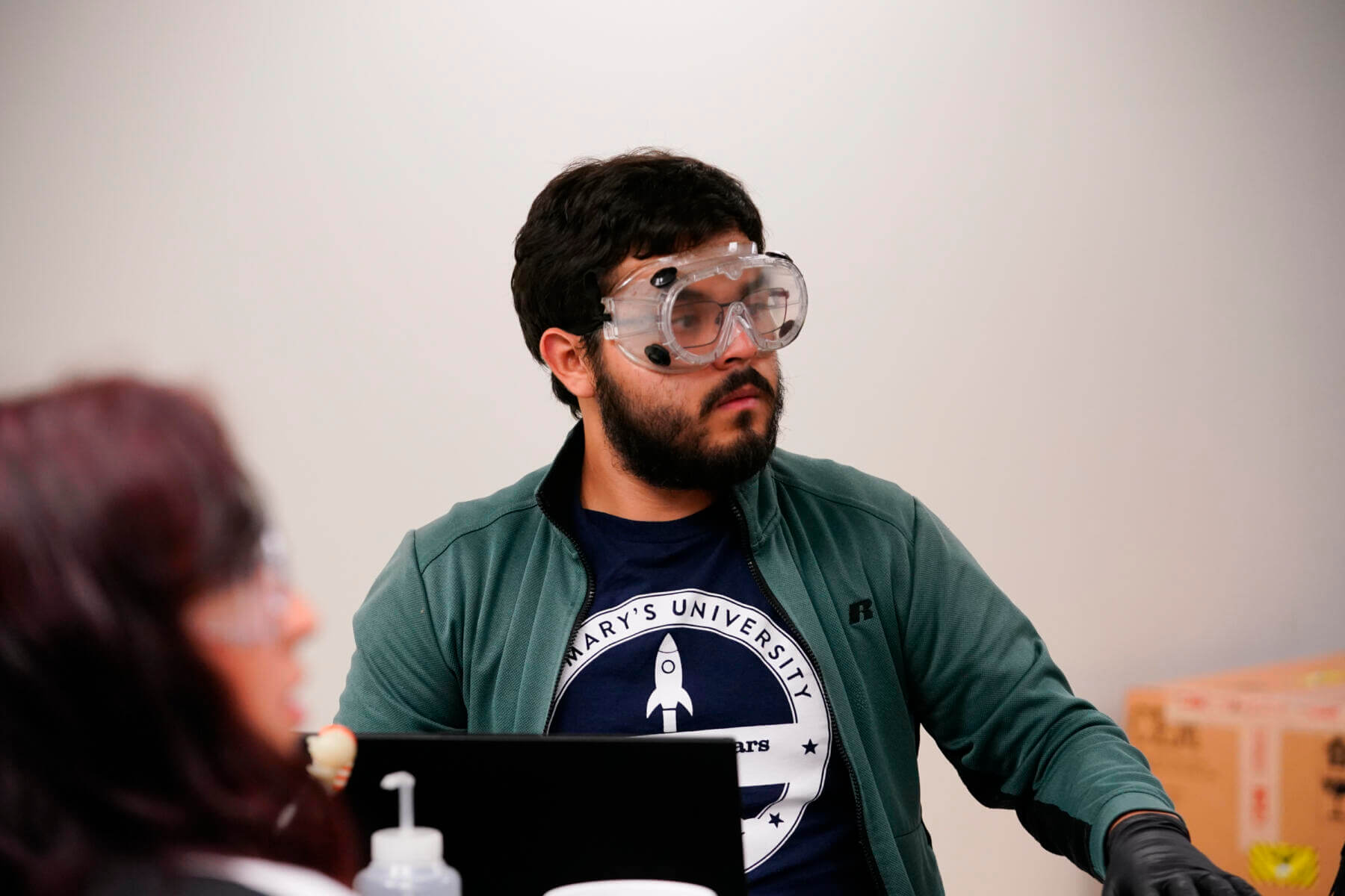 Male student wearing protective goggles looking to the side. In front of him is a laptop and science equipment. A female student wearing protective goggles turns to look in the same direction.