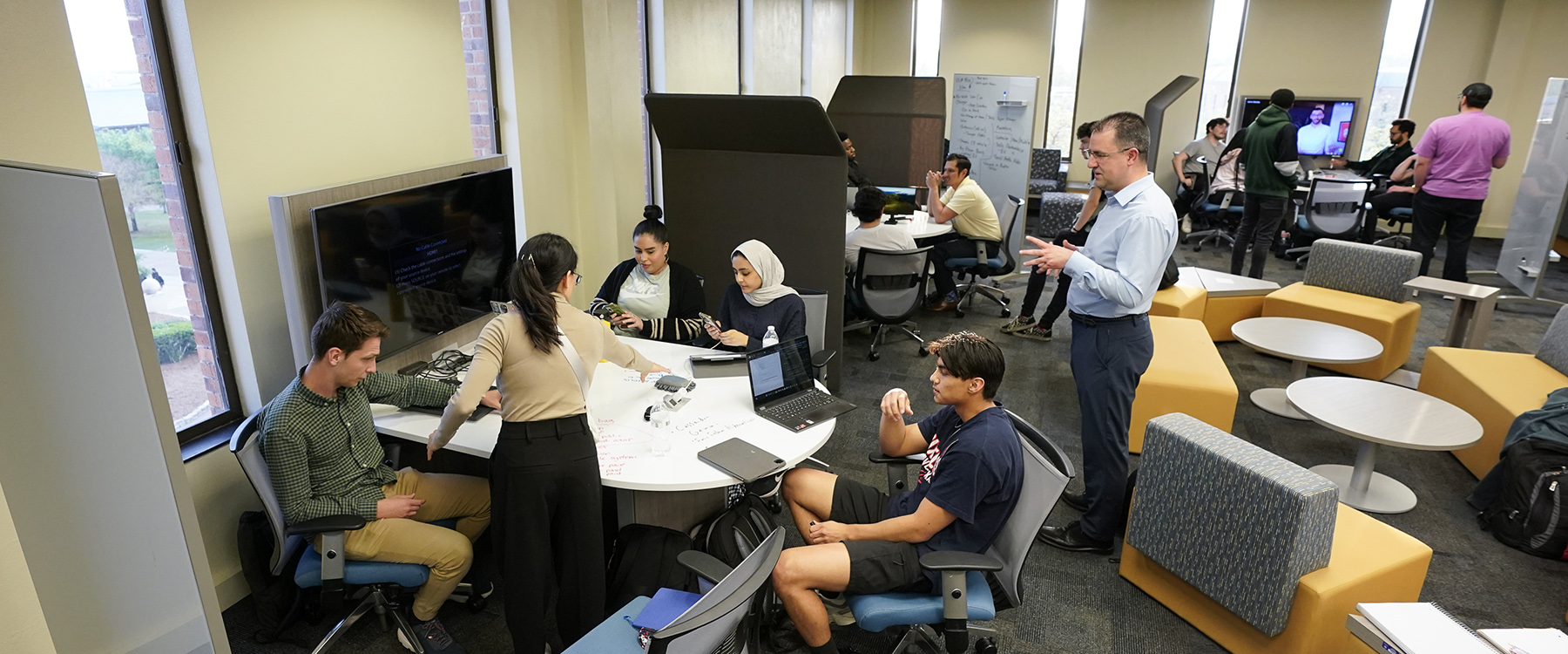 A group of students gather around a table discussing while a professor stands to the side watching them. 